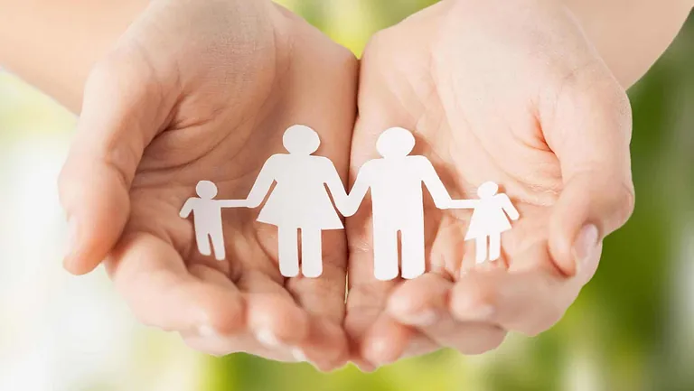 Hender som holder papirfigurer av en familie med to voksne og to barn mot en uskarp, grønn bakgrunn